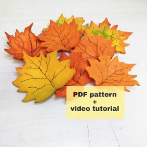 May include: A pile of felt maple leaves in shades of orange and yellow with black stitching outlining the veins. The leaves are arranged in a random pattern. The text "PDF pattern + video tutorial" is written in yellow on a white background.