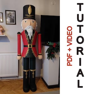 Könnte beinhalten: Eine große, dekorative Nussknacker-Figur. Sie hat einen schwarzen Hut mit goldenen Akzenten, eine rote Jacke mit goldenen Verzierungen und schwarze Stiefel. Der Text "TUTORIAL PDF + VIDEO" ist auf der rechten Seite zu sehen.