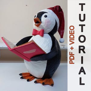 Può includere: Una statuetta di pinguino fatta a mano con un cappello da Babbo Natale e un papillon rossi, che tiene in mano un libro rosa. Il pinguino ha un piumaggio bianco e nero, zampe arancioni e un viso dettagliato. Il testo "PDF + VIDEO TUTORIAL" è incluso.