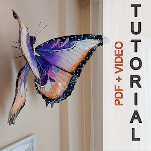 Peut inclure: Une sculpture de papillon colorée, avec des motifs complexes sur les ailes en violet, orange et blanc. Le papillon est fixé au mur. Le texte "TUTORIAL PDF + VIDEO" est visible sur le côté droit.