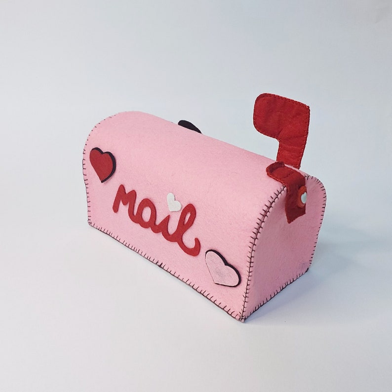 Pode incluir: Uma caixa de correio de feltro rosa com uma bandeira de feltro vermelha. A caixa de correio tem a palavra "mail" escrita em letras de feltro vermelho e &eacute; decorada com cora&ccedil;&otilde;es de feltro vermelho e branco.