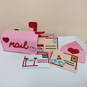 Pode incluir: Uma caixa de correio de feltro rosa com a palavra "mail" na frente, cercada por letras e envelopes de feltro. Os envelopes t&ecirc;m cora&ccedil;&otilde;es, p&aacute;ssaros e um calend&aacute;rio com o n&uacute;mero 14.