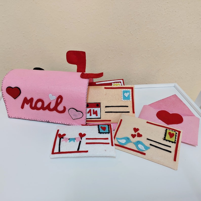 Pode incluir: Uma caixa de correio de feltro rosa com a palavra "mail" na frente, cercada por letras e envelopes de feltro. Os envelopes t&ecirc;m cora&ccedil;&otilde;es, p&aacute;ssaros e um calend&aacute;rio com o n&uacute;mero 14.