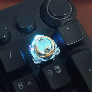 Może przedstawiać: Zbliżenie na świecący niebieski i złoty klawisz na czarnej klawiaturze. Klawisz jest niestandardowym wzorem z przezroczystą obudową z tworzywa sztucznego i złotą metalową ramką. Klawisz jest podświetlany od dołu, tworząc jasny niebieski blask.