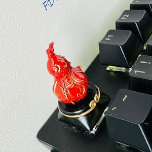 以下が含まれることがあります： 黒いキーボードの上に置かれた、炎の形をした赤と金の装飾キーキャップ。キーキャップには複雑な金の装飾が施されています。キーボードのキーは黒色です。「ETSY: ↓ F2AAAAA」という文字が青いインクで壁に書かれています。