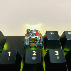 以下が含まれることがあります： 黒、グレー、オレンジのカラーリングが施された、小さく、詳細な機械式キーボードのキーキャップ。キーキャップは、1、2、3、4とラベル付けされたキーがついた黒い機械式キーボードの上に置かれている。