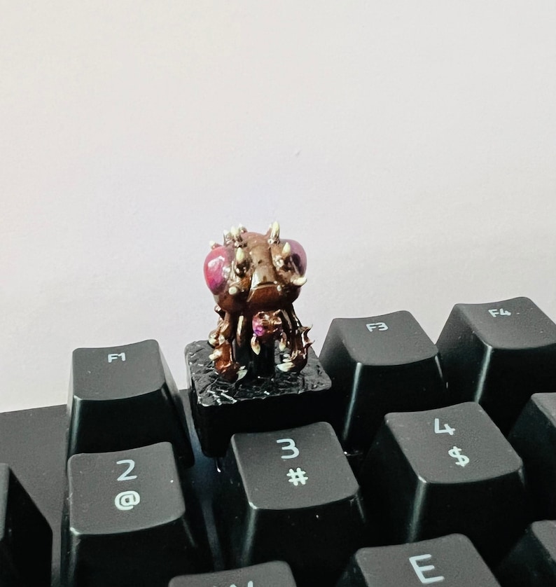 Puede incluir: Una criatura marr&oacute;n y negra con ojos rojos y p&uacute;as en la cabeza est&aacute; sentada sobre una tecla de teclado negra. La tecla est&aacute; etiquetada con el n&uacute;mero "3" y el s&iacute;mbolo "#"