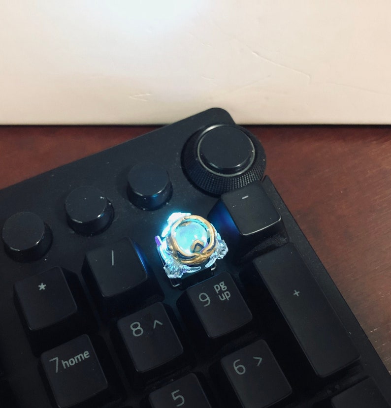 Może przedstawiać: Zbliżenie na świecący niebieski i złoty klawisz na czarnej klawiaturze. Klawisz jest niestandardowym wzorem z przezroczystą obudową z tworzywa sztucznego i złotą metalową ramką. Klawisz jest podświetlany od dołu, tworząc jasny niebieski blask.