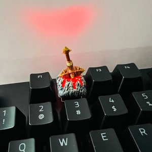 Op de afbeelding: Een gouden en zwarte miniatuur zwaard toetsenkap met een rood en wit ontwerp, dat op een zwart toetsenbord met witte toetsen zit, die met cijfers en letters zijn gelabeld.