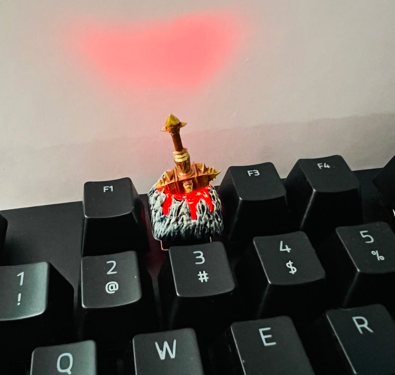 Op de afbeelding: Een gouden en zwarte miniatuur zwaard toetsenkap met een rood en wit ontwerp, dat op een zwart toetsenbord met witte toetsen zit, die met cijfers en letters zijn gelabeld.