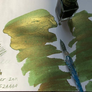 May include: A green and gold shimmer ink with gold glitter. The ink is in a glass bottle with a dropper. The ink is being used with a glass dip pen. The ink is labeled "R'lyeh shimmer ink shop: F2AAAA"
