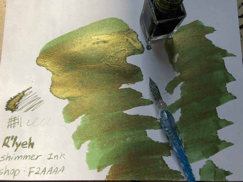 May include: A green and gold shimmer ink with gold glitter. The ink is in a glass bottle with a dropper. The ink is being used with a glass dip pen. The ink is labeled "R'lyeh shimmer ink shop: F2AAAA"