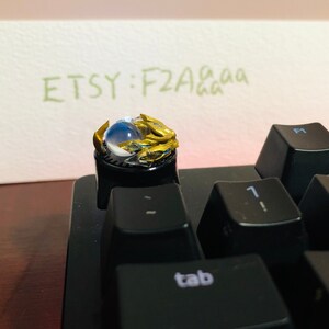 Puede incluir: Un tap&oacute;n de tecla dorado y azul con un orbe transparente en el centro. El tap&oacute;n de tecla est&aacute; en un teclado negro con la palabra "tab" en una de las teclas. El texto "ETSY: F2Aaaa" est&aacute; escrito en un papel en el fondo.