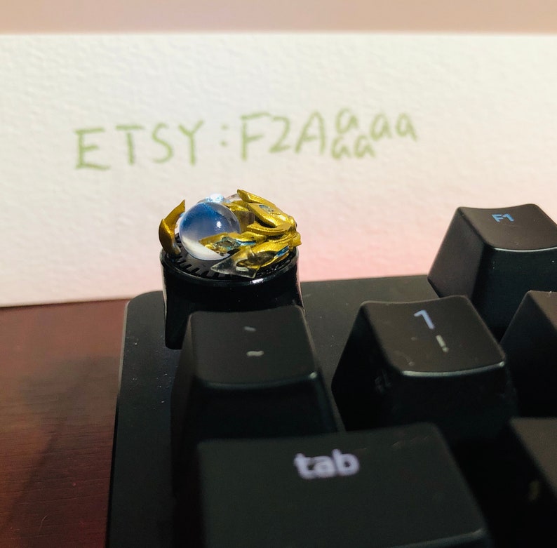 Puede incluir: Un tap&oacute;n de tecla dorado y azul con un orbe transparente en el centro. El tap&oacute;n de tecla est&aacute; en un teclado negro con la palabra "tab" en una de las teclas. El texto "ETSY: F2Aaaa" est&aacute; escrito en un papel en el fondo.