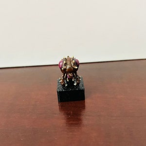 Puede incluir: Una peque&ntilde;a criatura marr&oacute;n similar a un insecto con patas espinosas y grandes ojos morados. La criatura est&aacute; de pie sobre una base negra.