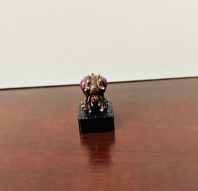 Puede incluir: Una peque&ntilde;a criatura marr&oacute;n similar a un insecto con patas espinosas y grandes ojos morados. La criatura est&aacute; de pie sobre una base negra.