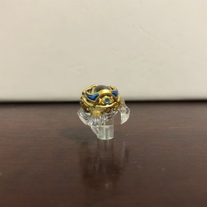 Może przedstawiać: Złoty i niebieski przedmiot dekoracyjny z przezroczystą szklaną kulą w środku. Przedmiot stoi na przezroczystym plastikowym stojaku.