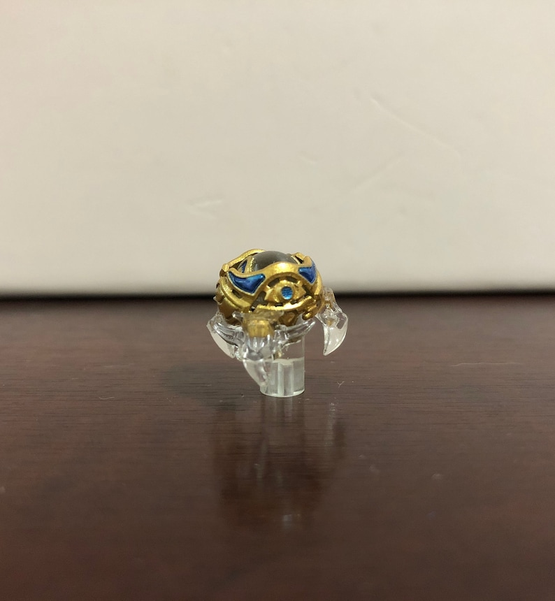 Może przedstawiać: Złoty i niebieski przedmiot dekoracyjny z przezroczystą szklaną kulą w środku. Przedmiot stoi na przezroczystym plastikowym stojaku.