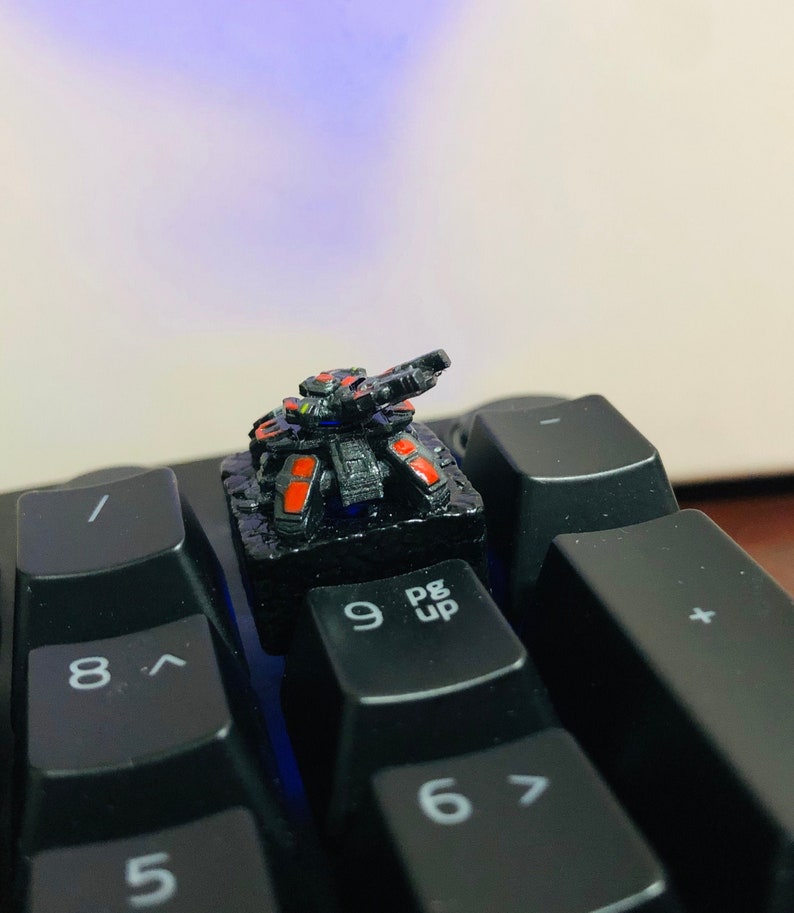 May include: A black and red miniature tank sits on a black keyboard with the keys '9', 'Pg Up', '6', and '8' visible. The tank has a small, raised gun turret and a detailed design.