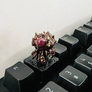 Puede incluir: Una peque&ntilde;a figura impresa en 3D, detallada, de una criatura con un cuerpo marr&oacute;n y espinoso, un ojo rojo y cuatro patas. La figura est&aacute; sentada sobre una tecla de teclado negra.