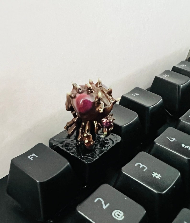 Puede incluir: Una peque&ntilde;a figura impresa en 3D, detallada, de una criatura con un cuerpo marr&oacute;n y espinoso, un ojo rojo y cuatro patas. La figura est&aacute; sentada sobre una tecla de teclado negra.