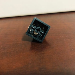 Puede incluir: Un keycap de teclado mec&aacute;nico cuadrado negro con un dise&ntilde;o en forma de cruz y un patr&oacute;n de engranaje.