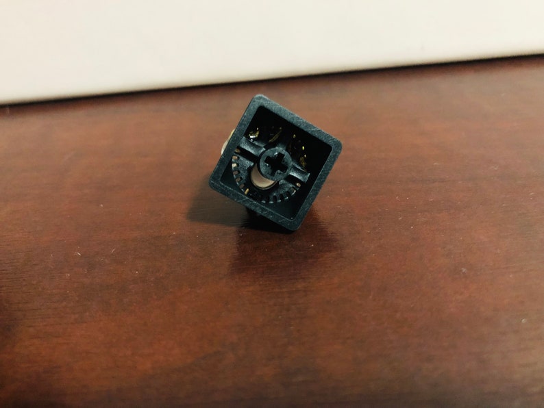Puede incluir: Un keycap de teclado mec&aacute;nico cuadrado negro con un dise&ntilde;o en forma de cruz y un patr&oacute;n de engranaje.