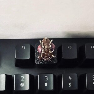 Puede incluir: Un teclado negro con una tecla personalizada que presenta una criatura detallada, impresa en 3D, de color marr&oacute;n y rosa, con p&uacute;as blancas. La criatura tiene ojos rojos y una superficie texturizada.