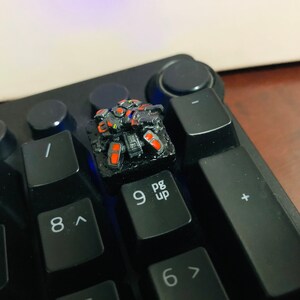 May include: A black keyboard with a custom keycap featuring a detailed, 3D printed robot design. The robot is painted in black, red, and yellow. The keycap is on the 'pg up' key.
