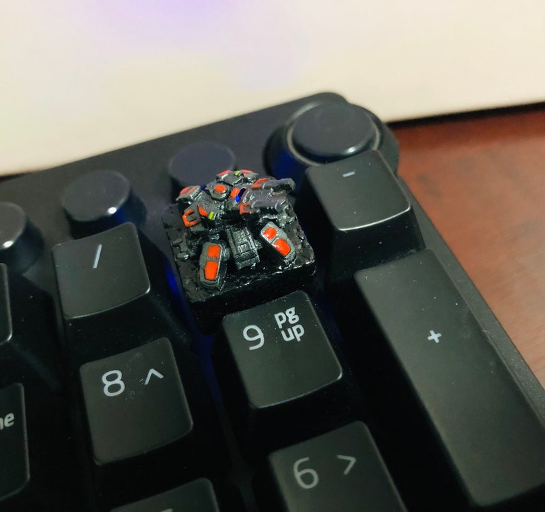 May include: A black keyboard with a custom keycap featuring a detailed, 3D printed robot design. The robot is painted in black, red, and yellow. The keycap is on the 'pg up' key.