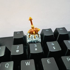 Op de afbeelding: Een gouden en bruine miniatuurfiguur van een krijger die een zwaard vasthoudt, zittend op een zwart toetsenbord met toetsen met de labels 1, 2, 3, 4, 5, Q, W, E, R, F1, F3 en F4.