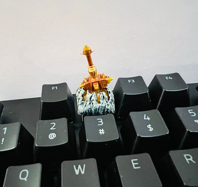 Op de afbeelding: Een gouden en bruine miniatuurfiguur van een krijger die een zwaard vasthoudt, zittend op een zwart toetsenbord met toetsen met de labels 1, 2, 3, 4, 5, Q, W, E, R, F1, F3 en F4.