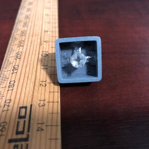 May include: A gray mechanical keyboard keycap with a clear plastic stem and a clear plastic housing. The keycap is designed to fit a standard mechanical keyboard.