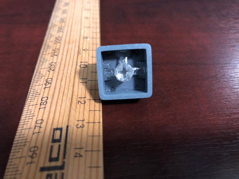 May include: A gray mechanical keyboard keycap with a clear plastic stem and a clear plastic housing. The keycap is designed to fit a standard mechanical keyboard.