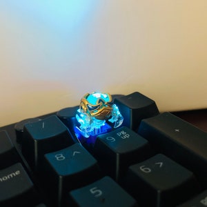 Może przedstawiać: Mały, złoty, świecący klawisz z niebieskim światłem emanującym z niego. Klawisz ma kształt kraba i znajduje się na czarnej klawiaturze z klawiszami oznaczonymi 7, 8, 9, 6, 5 i P/B Up.