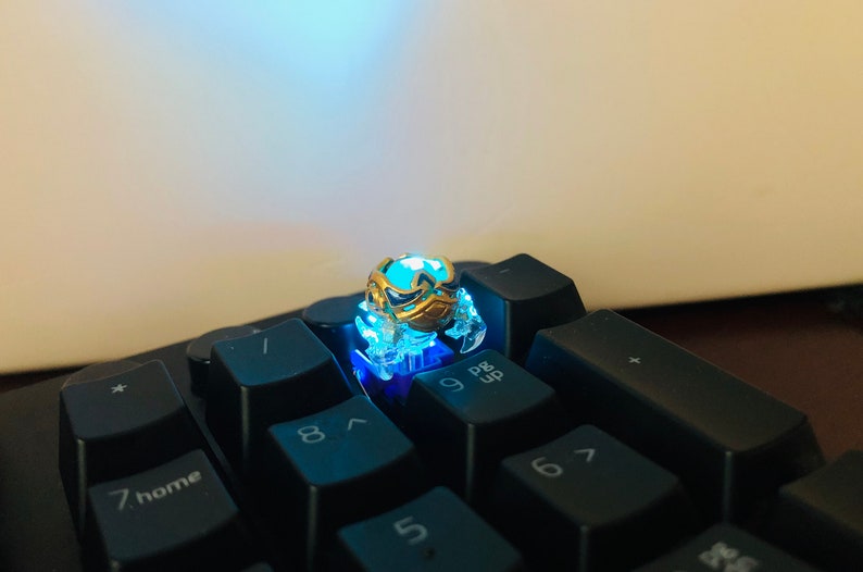 Może przedstawiać: Mały, złoty, świecący klawisz z niebieskim światłem emanującym z niego. Klawisz ma kształt kraba i znajduje się na czarnej klawiaturze z klawiszami oznaczonymi 7, 8, 9, 6, 5 i P/B Up.