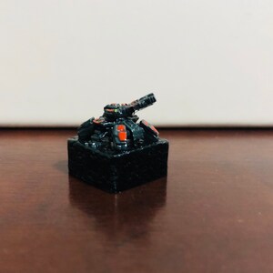 May include: A black and orange miniature tank with a raised gun turret on a black base.