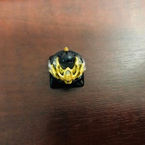 Puede incluir: Un keycap dorado y negro con un dise&ntilde;o detallado que presenta una gema negra central. El keycap est&aacute; dise&ntilde;ado para adaptarse a un teclado mec&aacute;nico.