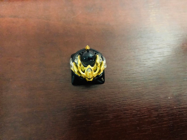 Puede incluir: Un keycap dorado y negro con un dise&ntilde;o detallado que presenta una gema negra central. El keycap est&aacute; dise&ntilde;ado para adaptarse a un teclado mec&aacute;nico.