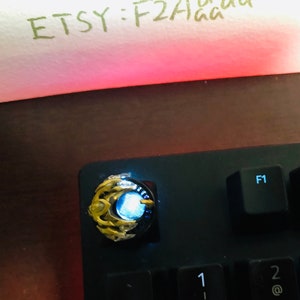 Puede incluir: Un tap&oacute;n de tecla dorado y azul con una luz azul brillante. El tap&oacute;n de tecla est&aacute; en un teclado negro con las teclas F1 y F2 visibles.