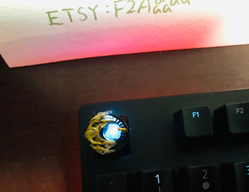 Puede incluir: Un tap&oacute;n de tecla dorado y azul con una luz azul brillante. El tap&oacute;n de tecla est&aacute; en un teclado negro con las teclas F1 y F2 visibles.