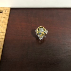 Może przedstawiać: Mały, przezroczysty szklany stojak na pierścionek z złotym i niebieskim pierścionkiem w środku. Pierścionek ma przezroczysty kamień w środku.