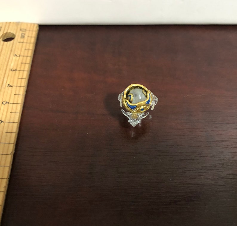 Może przedstawiać: Mały, przezroczysty szklany stojak na pierścionek z złotym i niebieskim pierścionkiem w środku. Pierścionek ma przezroczysty kamień w środku.