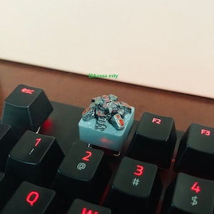 May include: A silver and red mechanical keyboard keycap featuring a detailed miniature robot. The keycap is sitting on a black keyboard with red backlighting.