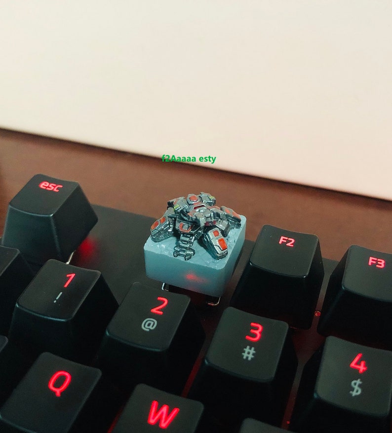 May include: A silver and red mechanical keyboard keycap featuring a detailed miniature robot. The keycap is sitting on a black keyboard with red backlighting.