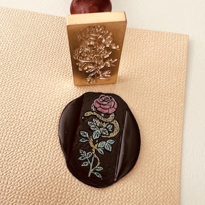 Könnte beinhalten: Ein Stempel aus Messing mit einem Design einer Schlange, die sich um eine Rose windet. Der Stempel wird verwendet, um ein Wachssiegel mit dem gleichen Design zu erstellen. Das Wachssiegel ist schwarz mit einer rosa Rose und einer grünen Schlange.