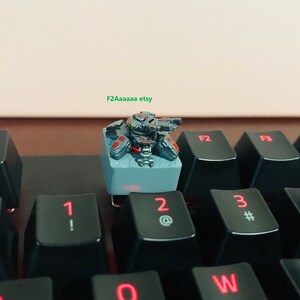 May include: A black and gray miniature robot figurine with red accents sits on a gray keycap. The keycap is on a black keyboard with red lettering. The robot has a gun and is in a fighting pose.