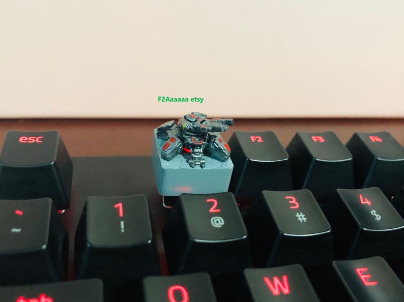 May include: A black and gray miniature robot figurine with red accents sits on a gray keycap. The keycap is on a black keyboard with red lettering. The robot has a gun and is in a fighting pose.