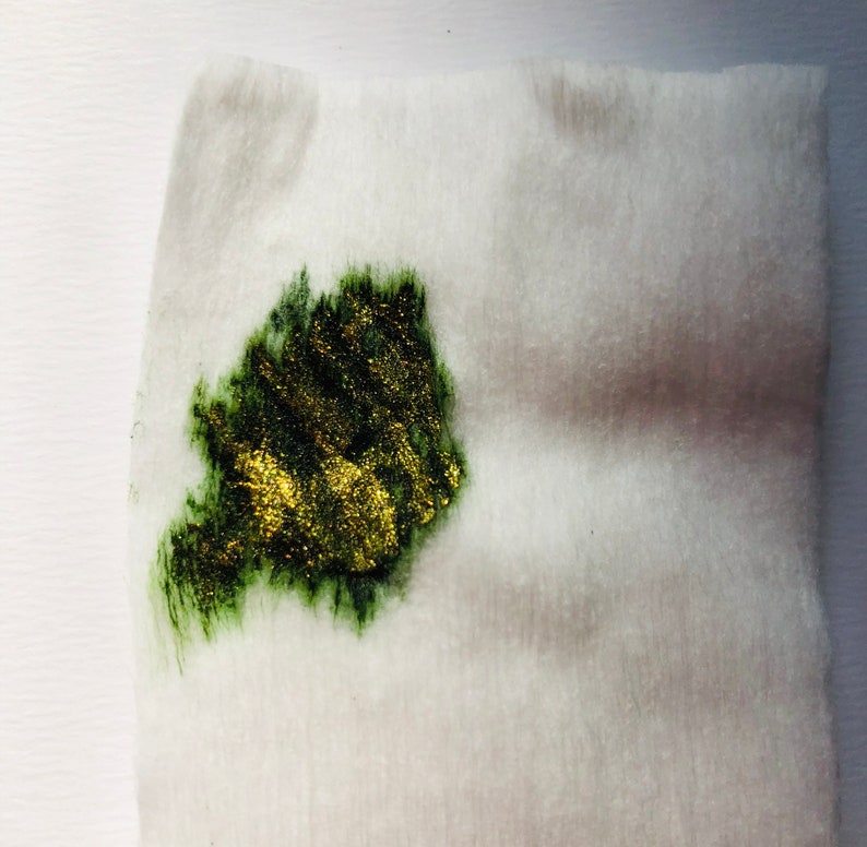 May include: A close-up of a white cotton pad with a green and gold splatter of paint on it.