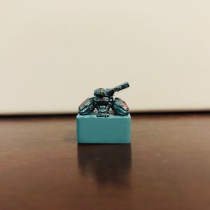 May include: A small, gray, and black miniature tank with a red stripe on the side, sitting on a gray base.